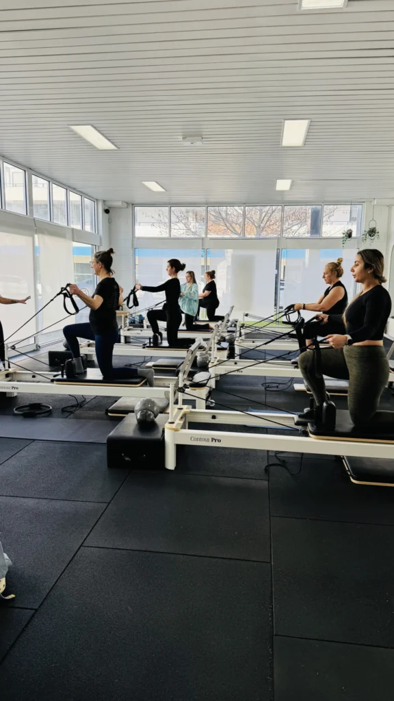 Group reformer Pilates class performing kneeling arm pulls with straps in a bright studio.