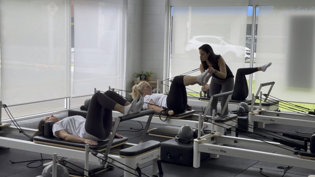 Pilates instructor assisting a client during a reformer class while participants perform leg exercises on the reformers.