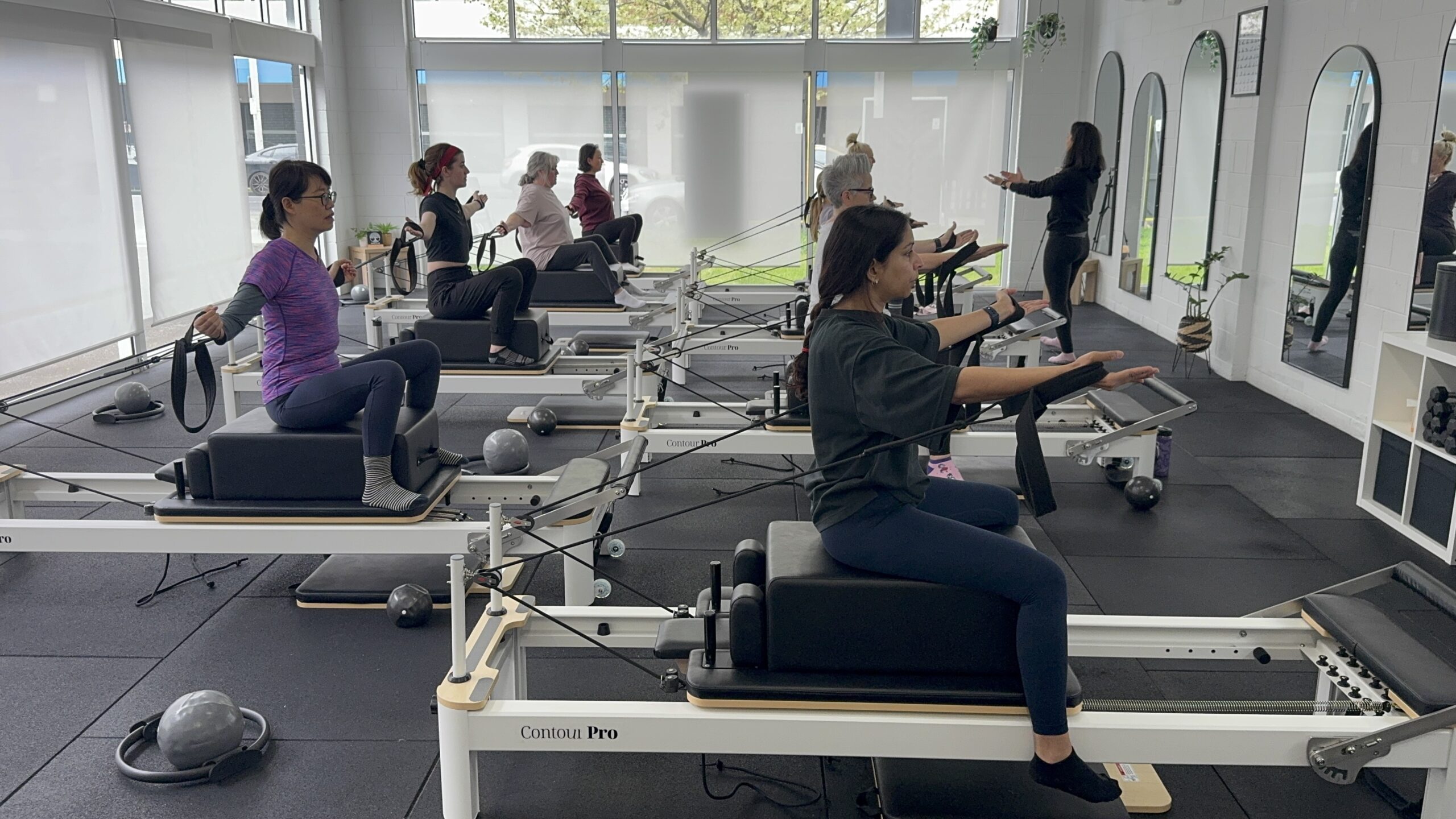 Group reformer Pilates class performing seated arm work with straps, guided by an instructor in the studio.