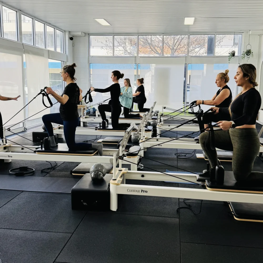 Group reformer Pilates class performing kneeling arm pulls with straps in a bright studio.